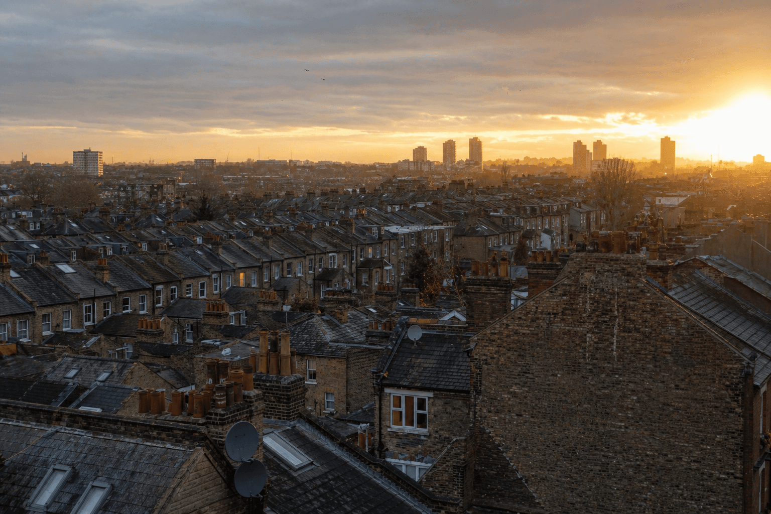 Wide view across UK terraced rooftops at golden hour, brick chimneys and slate roofs stretching to the horizon.