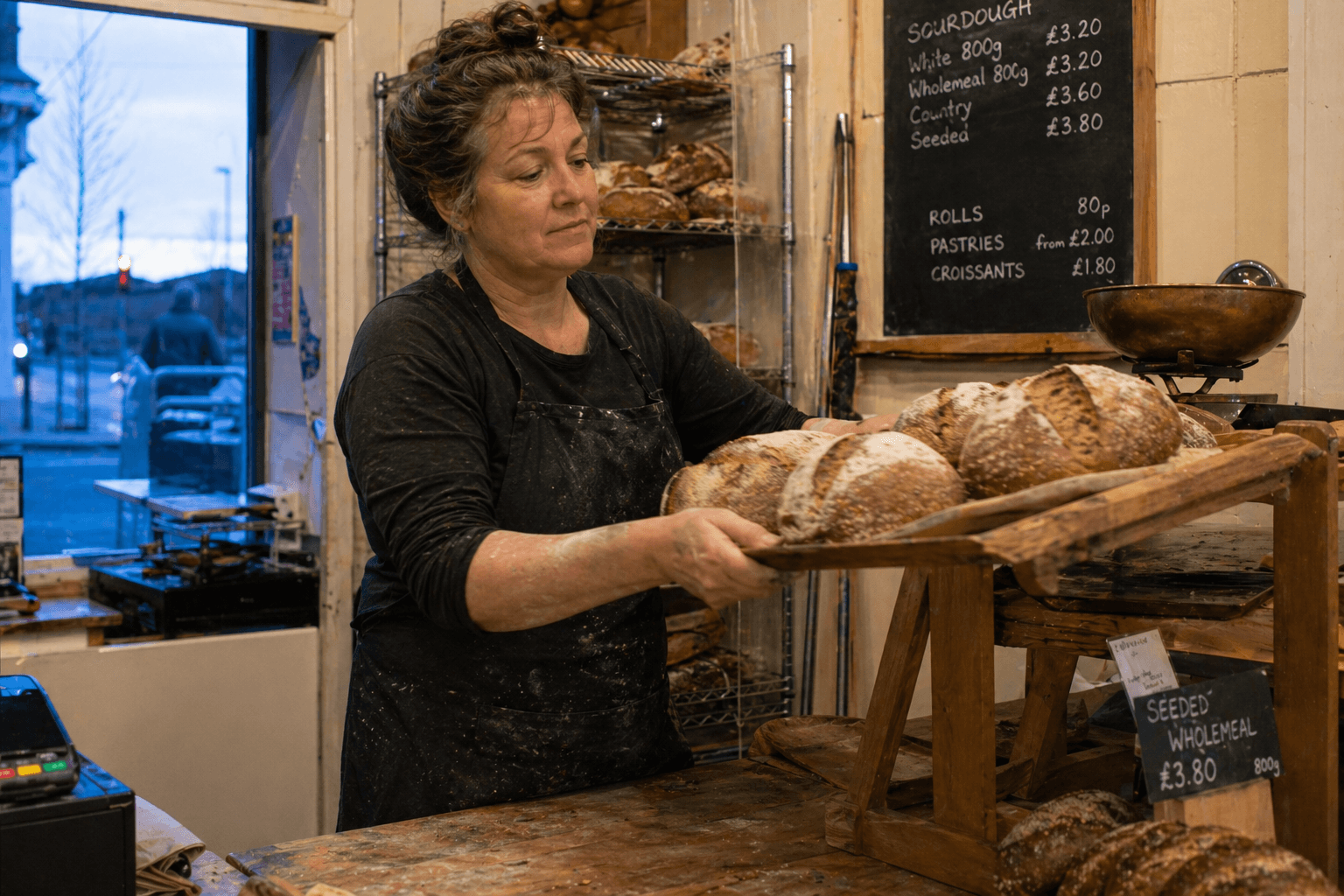 A baker in a flour-dusted apron placing a tray of fresh sourdough loaves onto a wooden display rack.